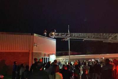 Mr. Brock receiving the pizza from the fire fighter. A crowd below on the ground is cheering.